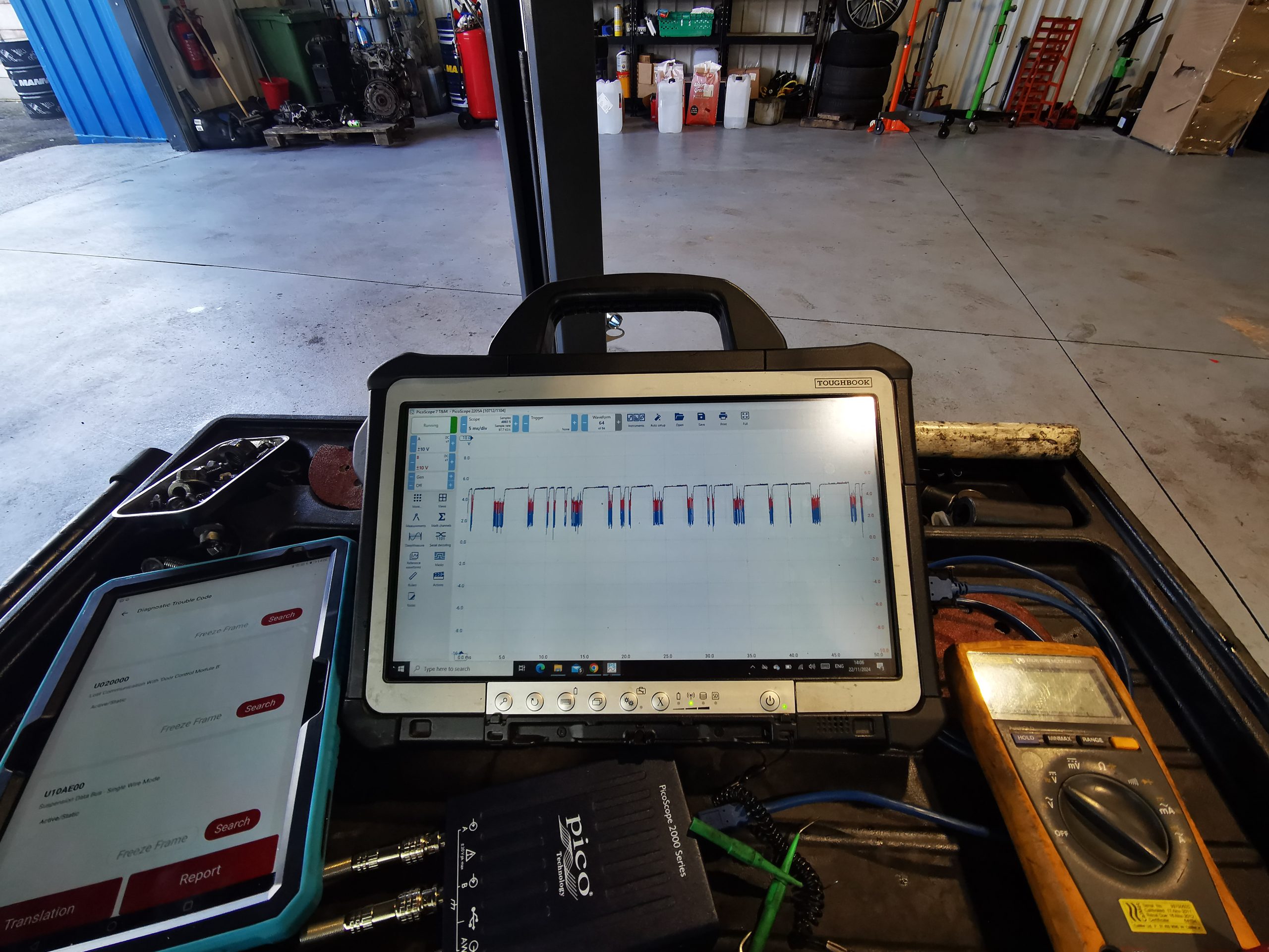 Automotive diagnostic equipment in a garage including a rugged laptop showing oscilloscope waveforms, a PicoScope, a multimeter and a scan tool tablet